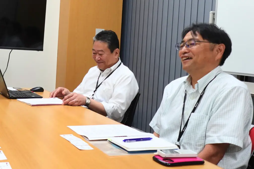 会議室で担当者2名が笑顔で談笑する様子。化学物質情報管理システムの運用手応えや効果について和やかに意見交換。