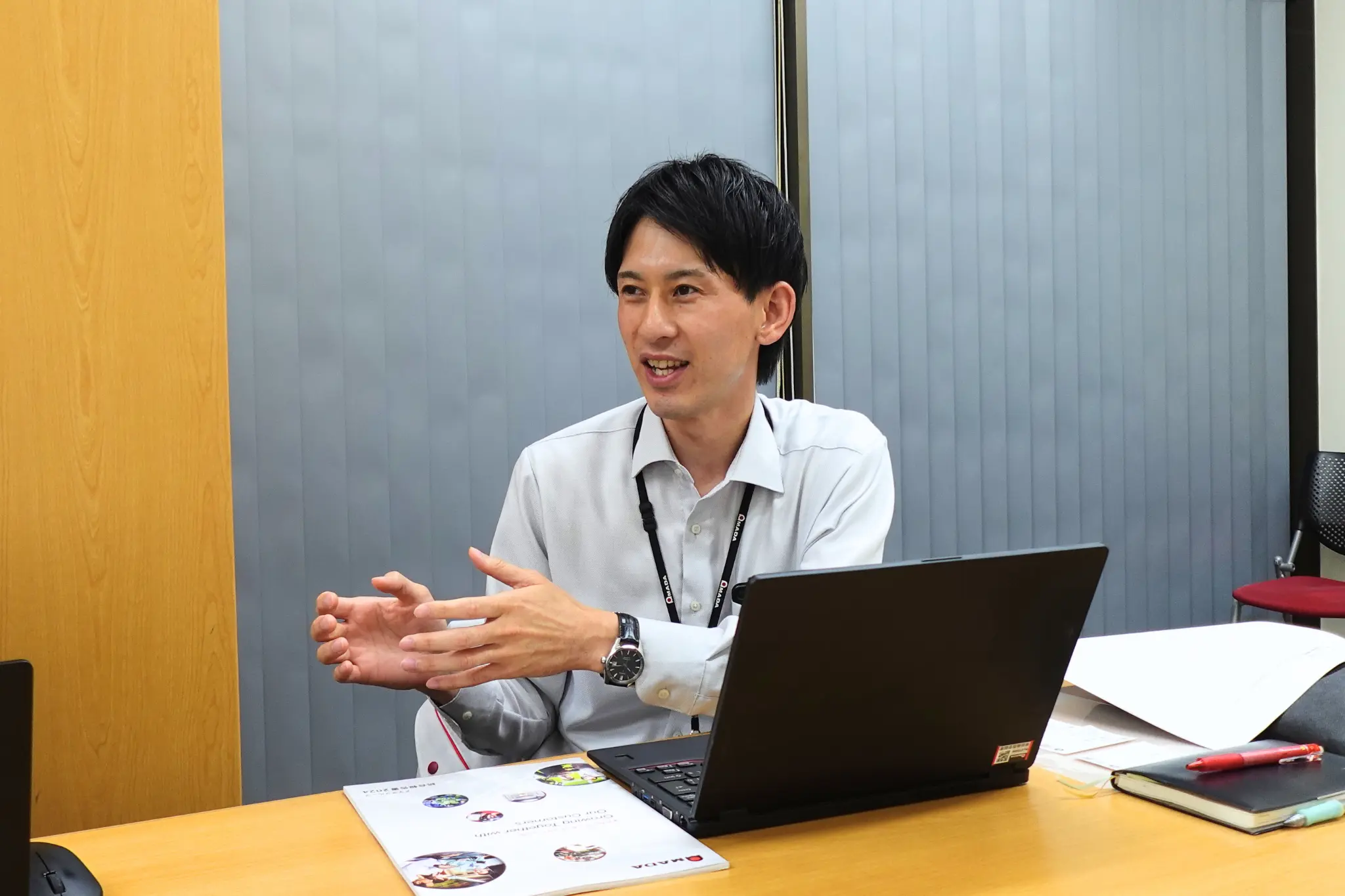 ノートPC前で担当者が手振りで説明する場面。現場での使い勝手やデータ検索性向上の実感を語るインタビュー。