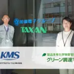 含有化学物質管理システム「グリーン調達マイスター」導入事例の集合写真。加賀電子グループ TAXAN の担当者と KMS コンサルタントが社屋前に並び、バナーに製品ロゴと会社ロゴが表示されている