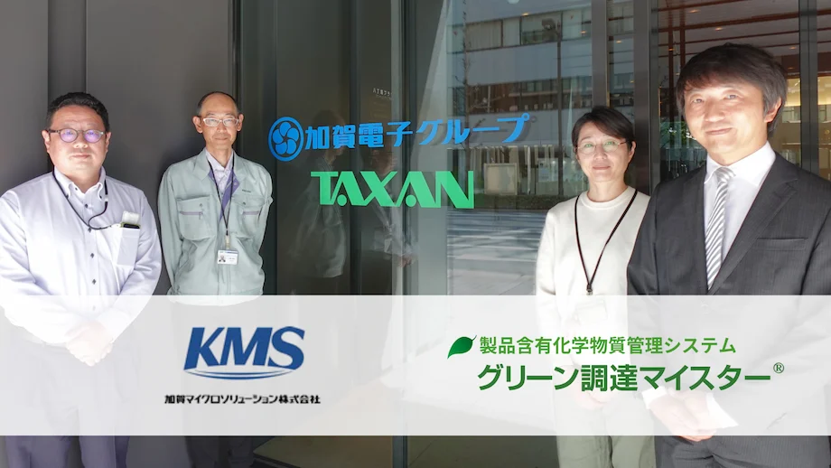含有化学物質管理システム「グリーン調達マイスター」導入事例の集合写真。加賀電子グループ TAXAN の担当者と KMS コンサルタントが社屋前に並び、バナーに製品ロゴと会社ロゴが表示されている