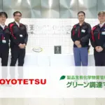 含有化学物質管理システム「グリーン調達マイスター」導入事例の集合写真。豊田鐵工（TOYOTETSU）の担当者とコンサルタントが社内ロビーに並び、バナーに TOYOTETSU ロゴと製品名が表示されている