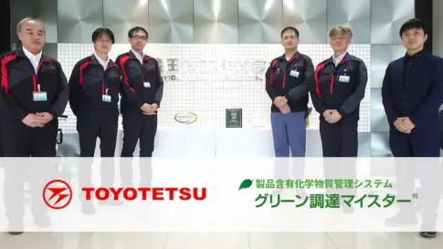 含有化学物質管理システム「グリーン調達マイスター」導入事例の集合写真。豊田鐵工（TOYOTETSU）の担当者とコンサルタントが社内ロビーに並び、バナーに TOYOTETSU ロゴと製品名が表示されている