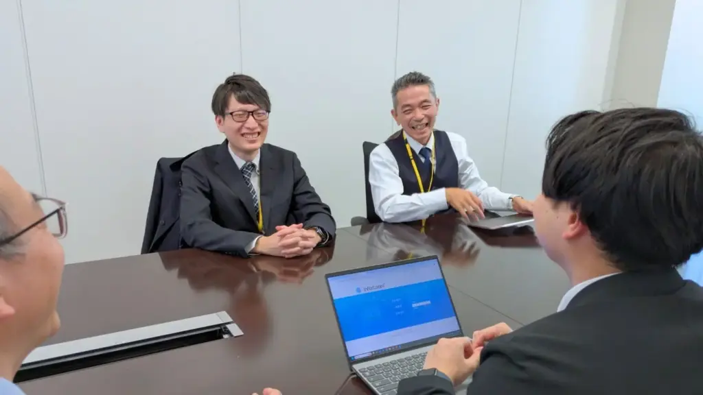 InforLoomのログイン画面を表示したノートPCを囲み、ユニプレス株式会社、UEL株式会社の担当者4名が笑顔で打ち合わせしている会議風景の写真