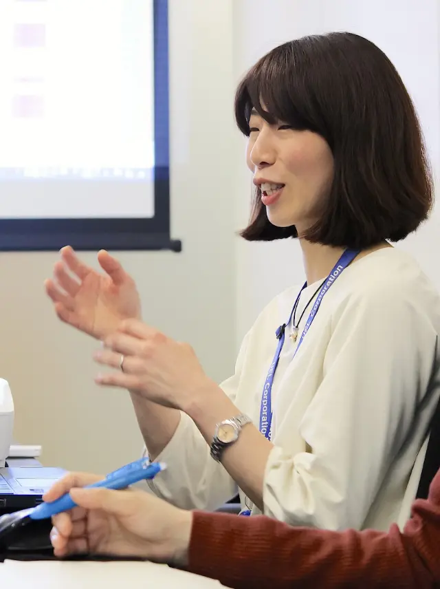 会議室で身振り手振りを交えて説明する木村さん。隣に同僚の手が写り、議論の様子が伝わる。