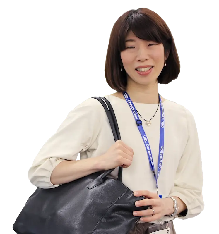 退勤シーンを想起させるカット。黒いバッグを肩にかけ、笑顔でこちらを向く木村さんの全身。