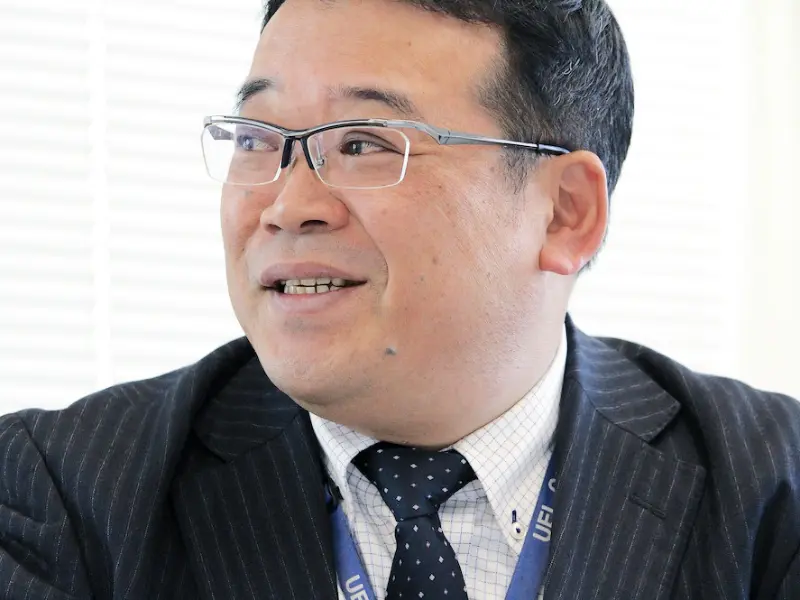 ブラインド越しの光を受けながら微笑む坂本さんのバストショット。穏やかな表情が印象的。