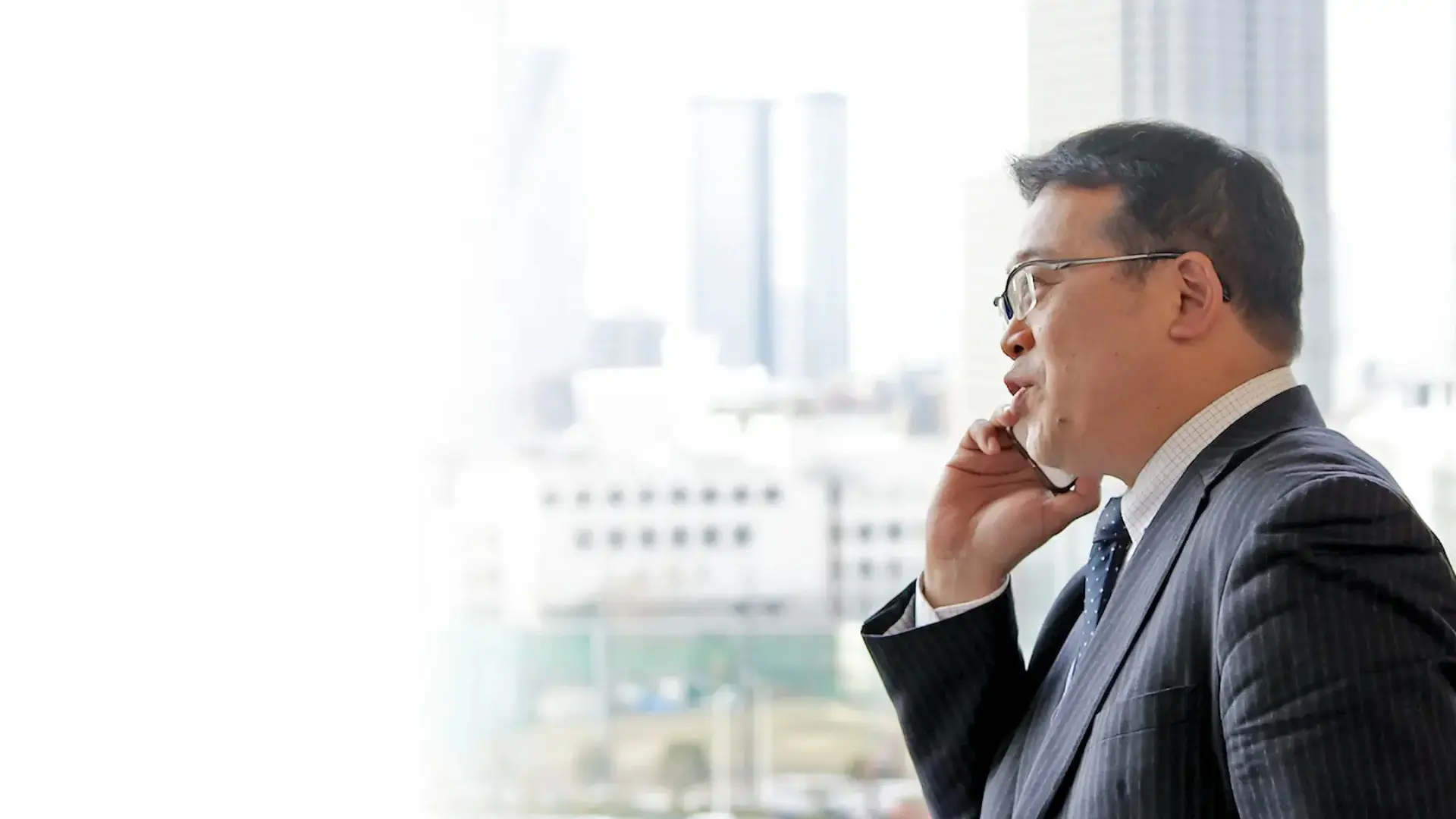 大きな窓辺で電話を耳に当てる坂本さん。背景に高層ビルがぼけて写り、ビジネスシーンを印象づけるメインビジュアル。