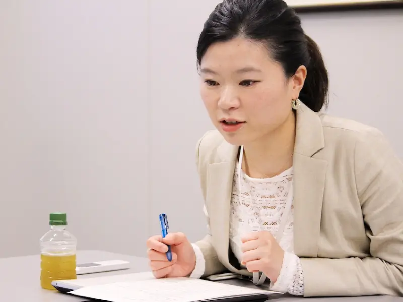 メモを取りつつ説明する住吉さん。ペットボトルとペンが映り、打合せの様子が分かる。