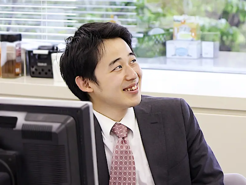 デスク前で笑顔を見せる丸山さんの仕事風景、窓外の緑が背景に映るインタビューショット。