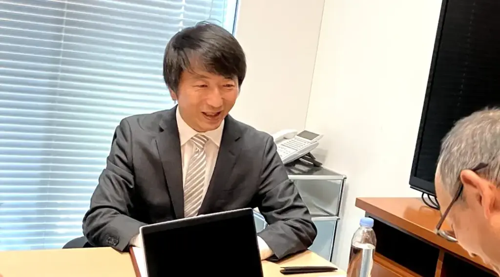 UELの営業担当者が商談テーブルで笑顔で説明し、手前には加賀マイクロソリューション担当者の頭部が写る商談シーン。